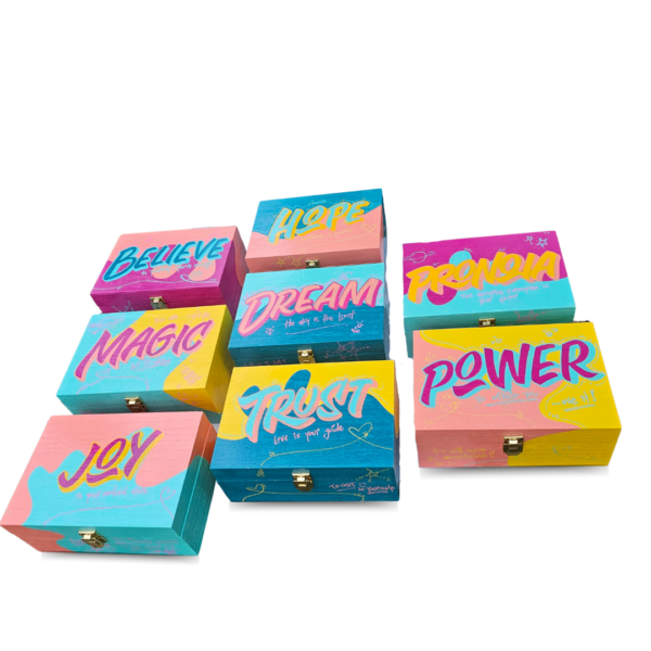 A collection of hand-painted wooden boxes with vibrant lettering featuring words like "Believe," "Hope," "Magic," "Dream," "Trust," "Pronoia," and "Power."